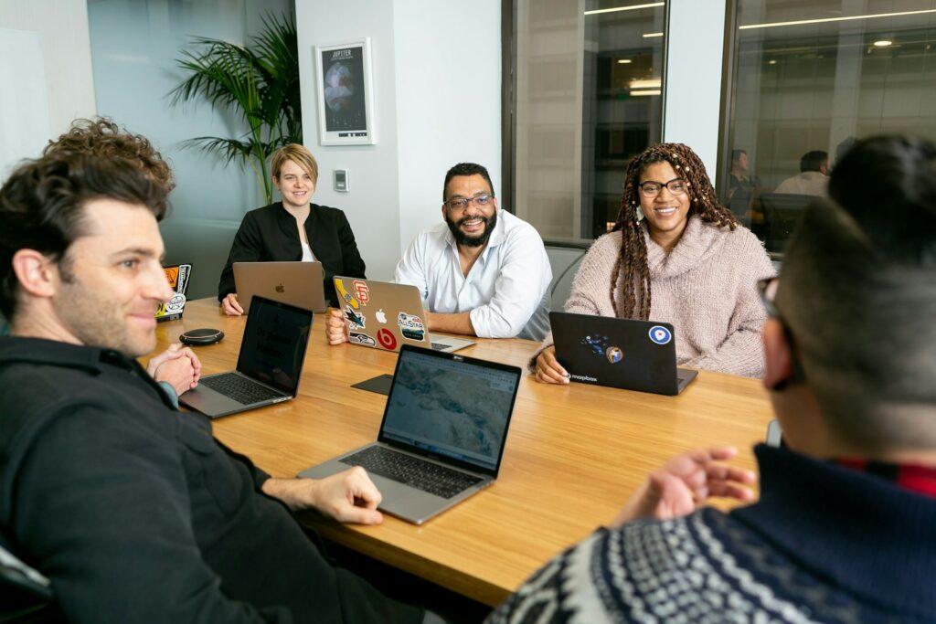 How to Work in Tech four people all on laptops, two men and two women, listen to person talking in a board meeting 